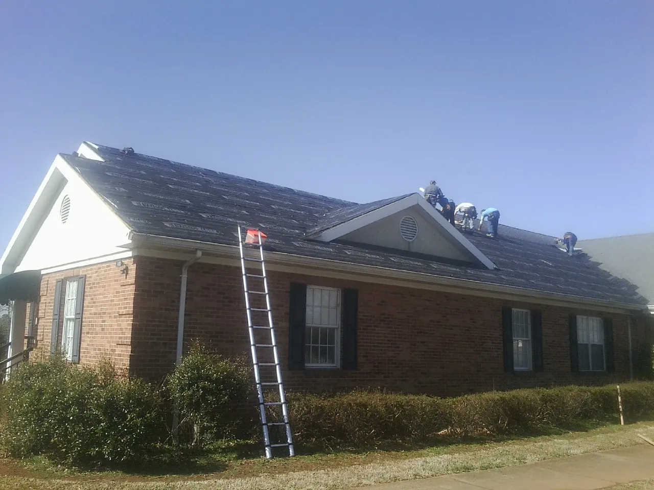 Crew tearing shingles off a brick ranch roof with a ladder set — Emergency Roof Repair work by our crew in Laurens