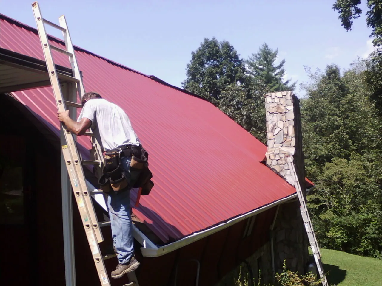 Roofer on a ladder beside a red metal roof and stone chimney — Soffit & Fascia Repair by Roofing Brothers in Laurens